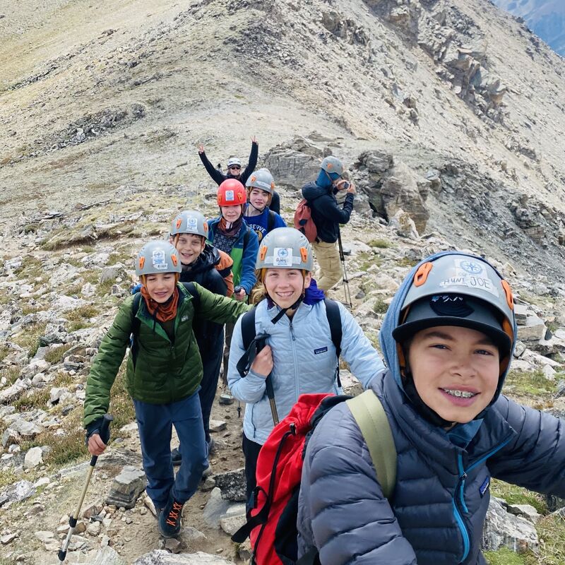 A group of young hikers are ascending a rocky mountain trail. They are wearing helmets and backpacks, suggesting they are prepared for a challenging climb. The landscape is barren and mountainous, with a steep incline. The hikers appear to be enjoying the adventure, with some smiling and looking at the camera. The overall impression is one of outdoor activity and teamwork in a rugged environment.
